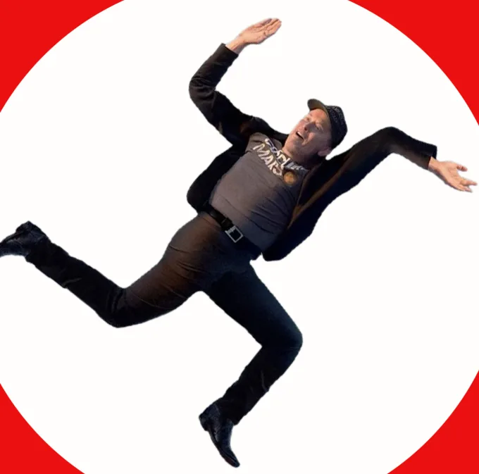 A man in a black jacket, grey t-shirt, black pants, and a cap is captured mid-air in a dramatic, falling pose against a…