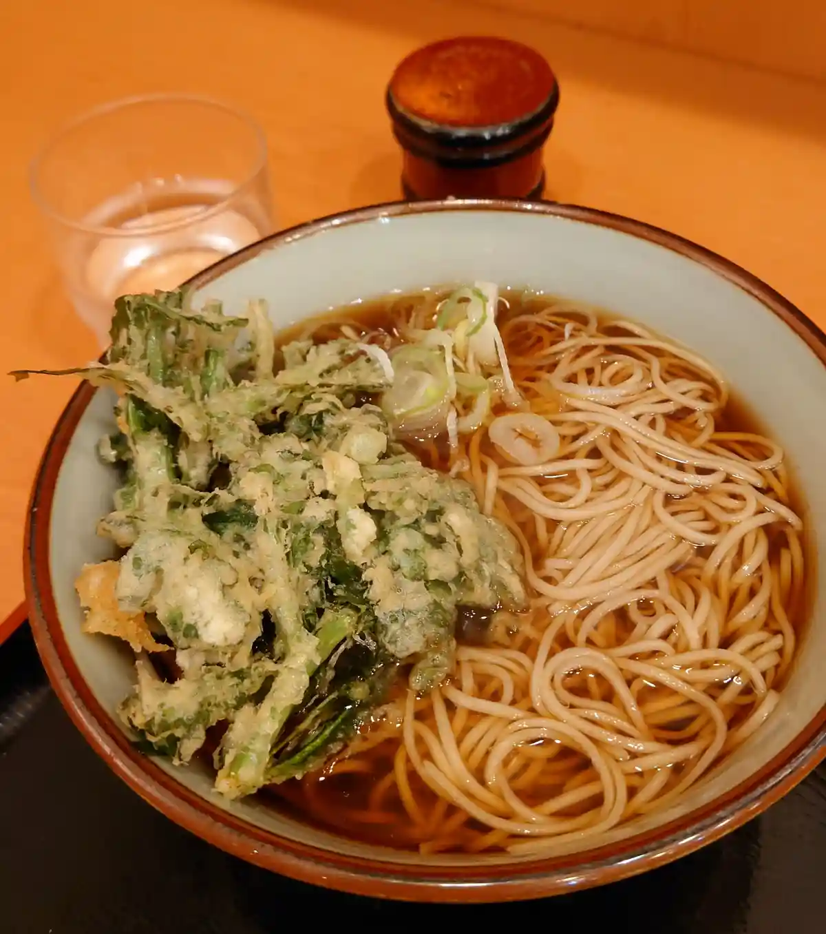 a close-up photo of a bowl of ramen with noodles in broth, topped with a large serving of tempura-fried green vegetables…