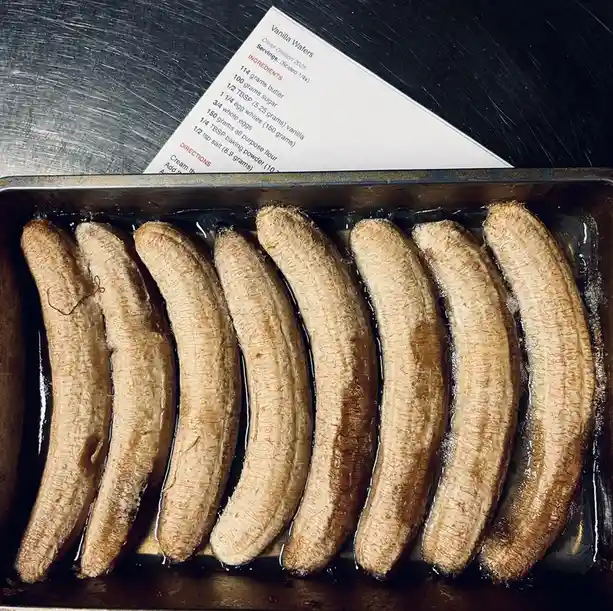 A metal baking tray holds seven peeled bananas submerged in a dark liquid, resembling a cooking mishap. Behind them, a…