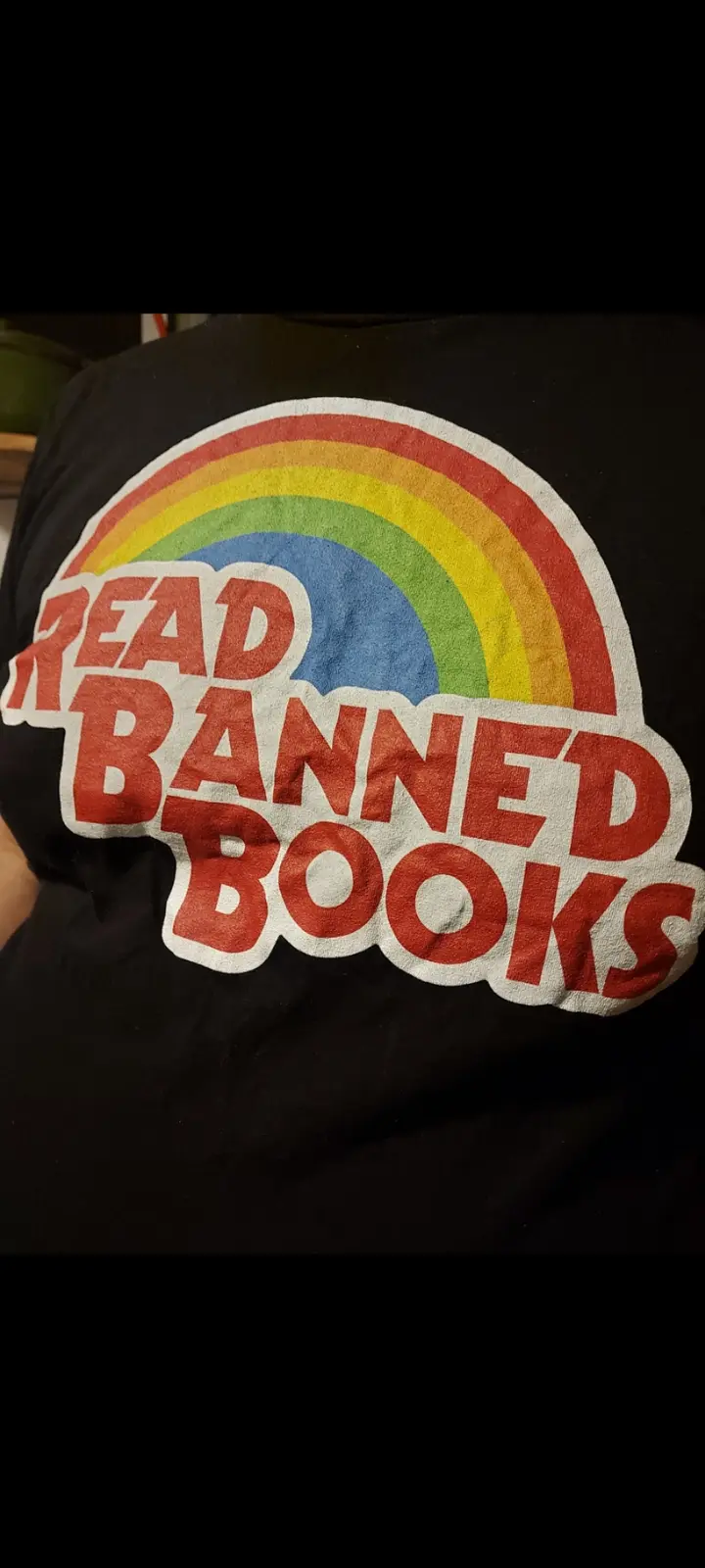 A close-up of a black t-shirt featuring a colorful rainbow arching over the bold red text 'READ BANNED BOOKS' with a white…