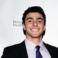A man in a suit and tie smiles broadly at the camera against a backdrop that reads 'Philadelphia Museum'. Overlaid text says…