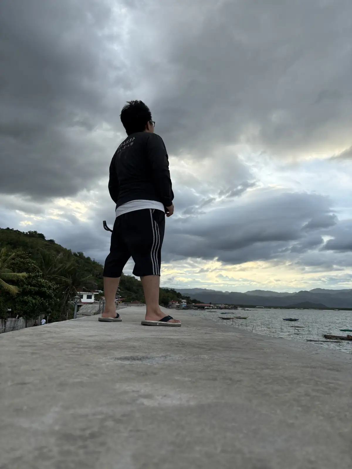 A person stands on a concrete pier facing a cloudy sky and water with boats, wearing a black long-sleeved shirt, black…