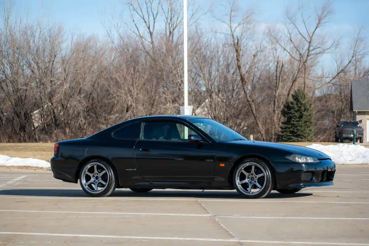A black Nissan Skyline R34 GTR is parked in a paved lot with snow patches and bare trees in the background. The car has…