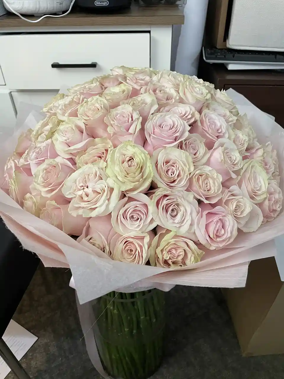 A large, round bouquet of light pink and cream-colored roses wrapped in matching tissue paper sits in a clear vase on a dark…