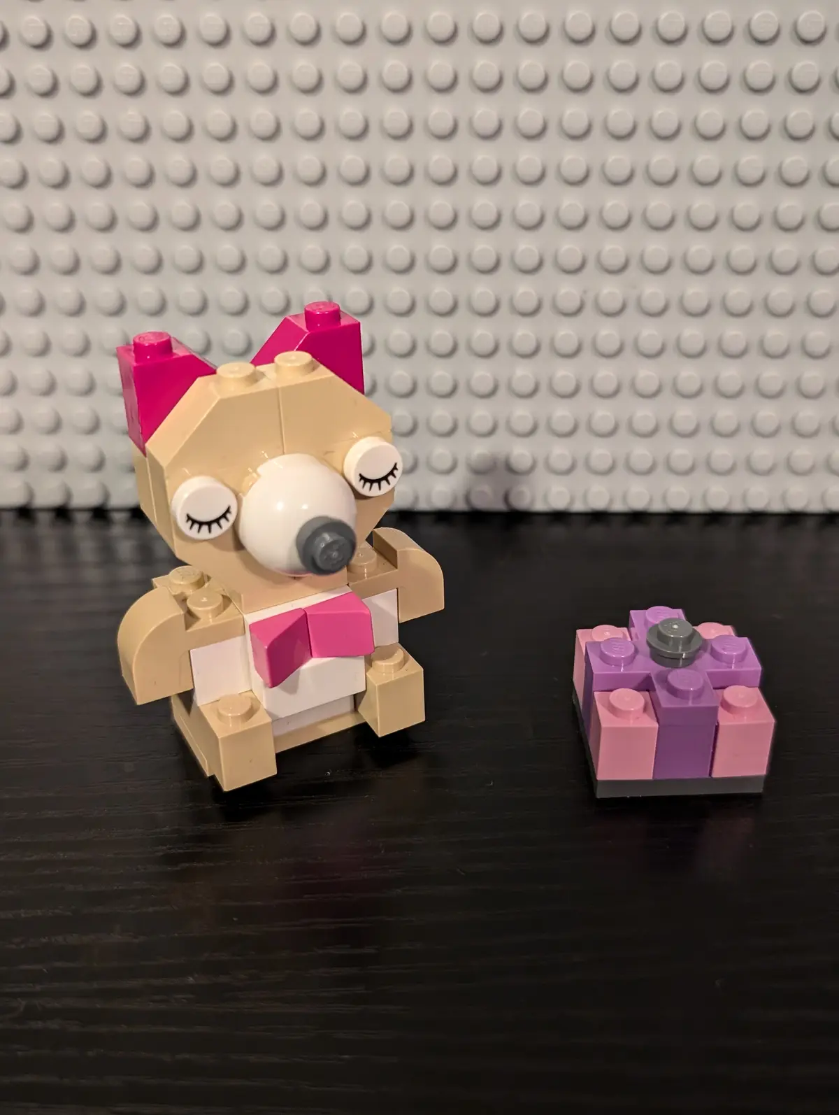 A small, tan LEGO figure with pink bow-like ears and closed eyes sits on a dark surface, looking at a small, purple and pink…
