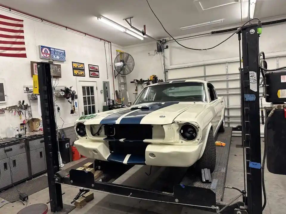 A white classic Ford Mustang with blue racing stripes is elevated on a car lift in a well-equipped garage. The front bumper…