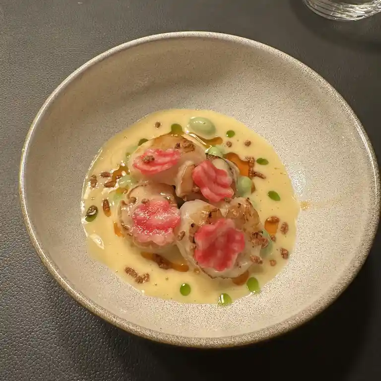 A top-down photo of a gourmet dish in a speckled ceramic bowl, featuring seared scallops in a creamy sauce, garnished with…