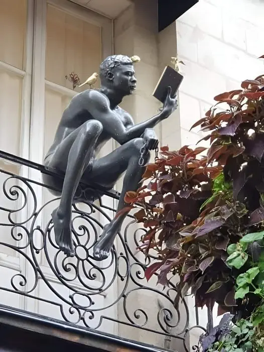A bronze statue of a nude man sits on an ornate wrought-iron balcony railing, holding an open book as if reading. Two small…