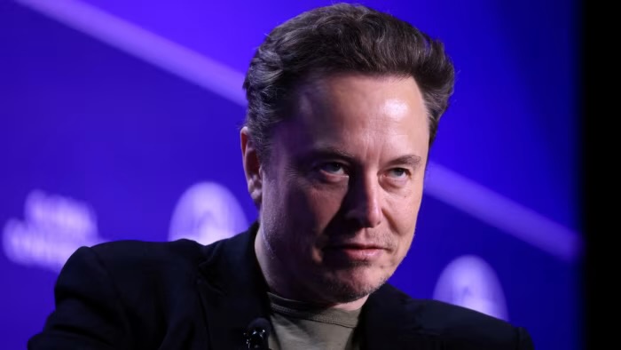 A close-up photo of Elon Musk looking slightly smug or amused, wearing a black jacket over a green shirt, against a blue…