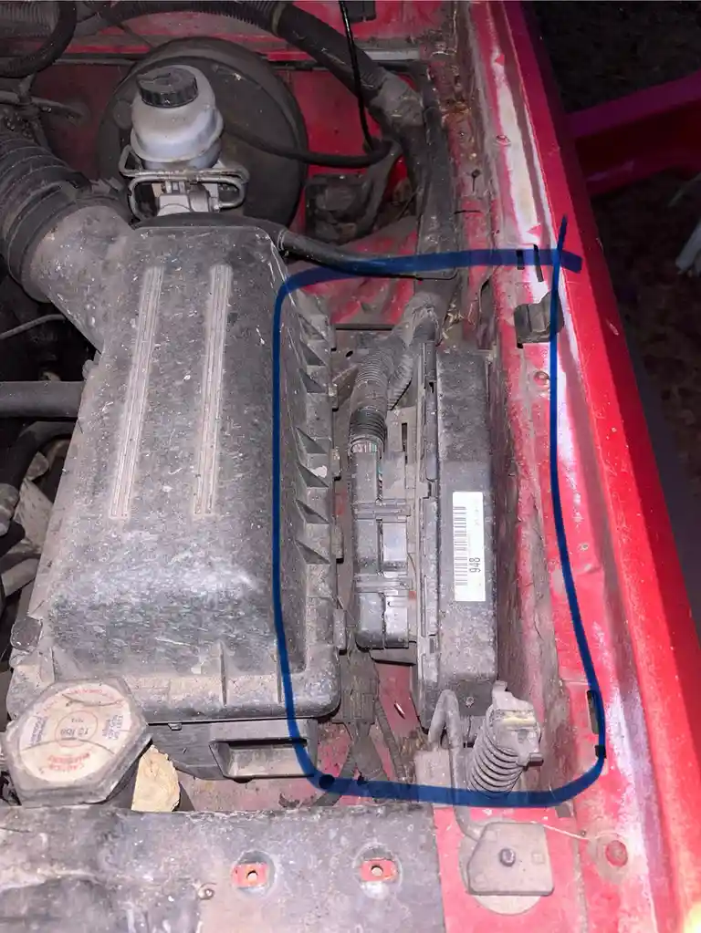 a close-up photo of a dusty red car engine bay shows a black plastic component outlined in blue, situated between the engine…