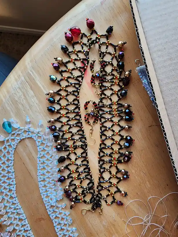 A close-up photo shows two intricate, beaded earrings shaped like spider webs, featuring black, gold, and red beads with…