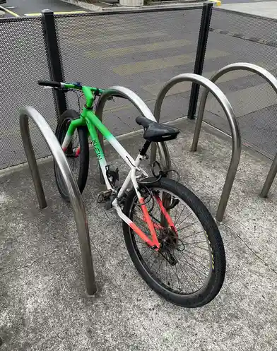 A bicycle with a green-to-white-to-orange gradient frame is locked to a metal bike rack. The bike is positioned at an angle…