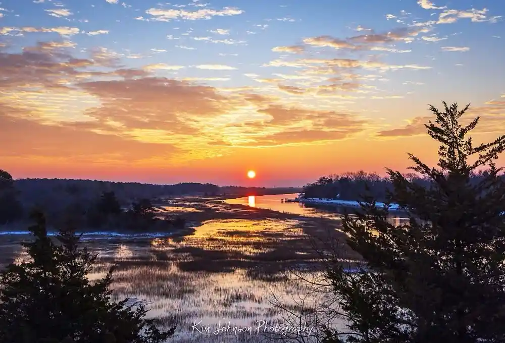 A serene landscape photograph captures a vibrant sunset over a wide river, with the sun low on the horizon casting golden…