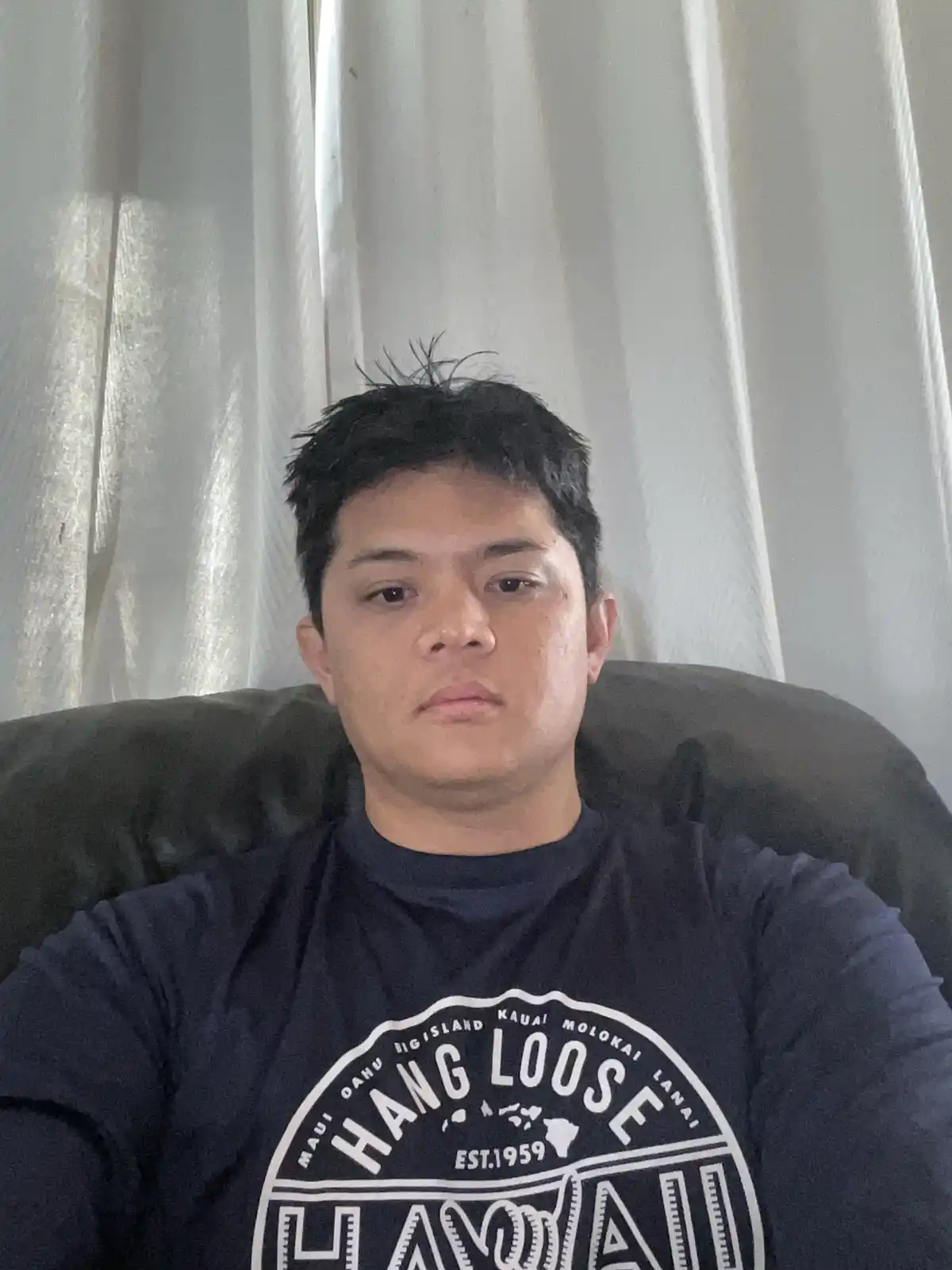 A person with short dark hair sits in a dark armchair in front of sheer white curtains, wearing a dark blue t-shirt with a…