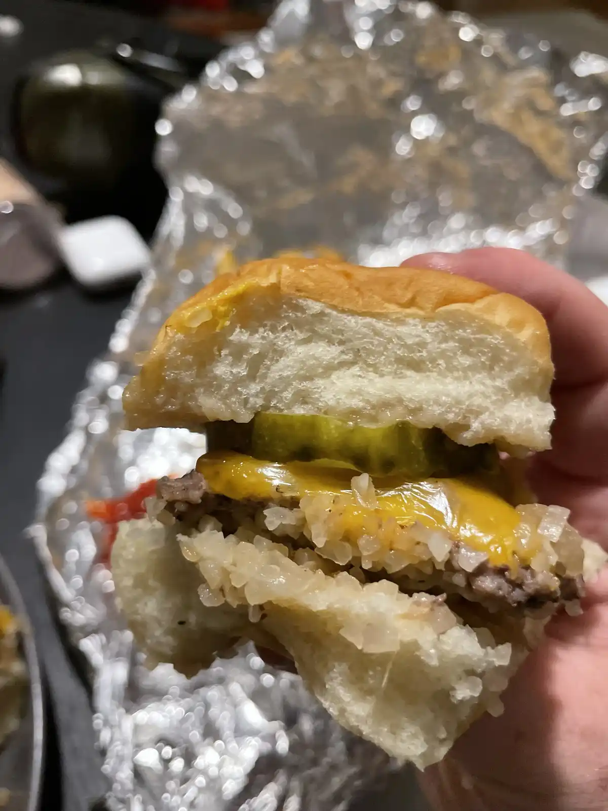 A hand holds a partially eaten burger with a visible pickle, melted cheese, and a beef patty, all nestled in a soft bun. The…
