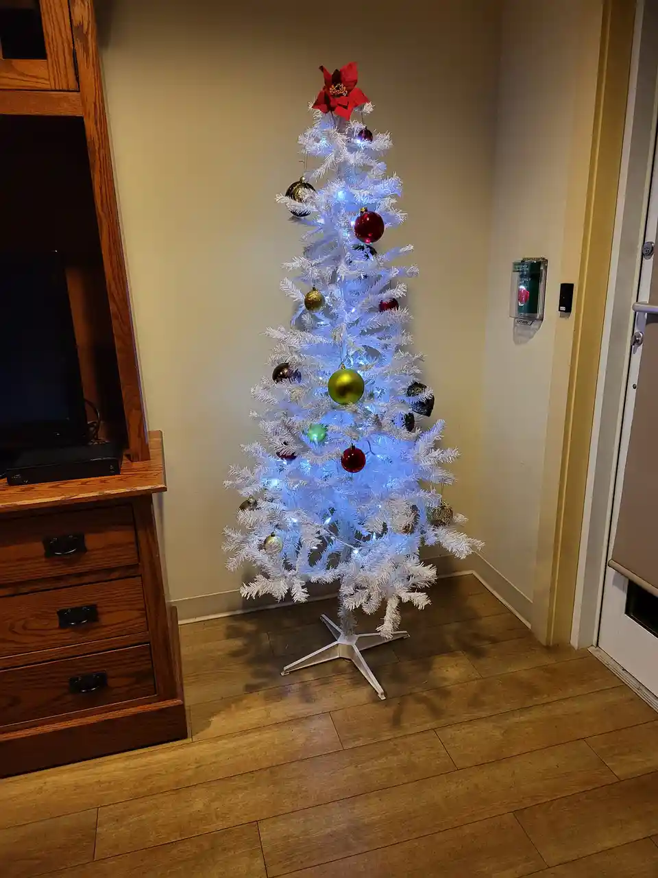 A white artificial Christmas tree decorated with blue lights, red and gold ornaments, and a red poinsettia topper stands in…