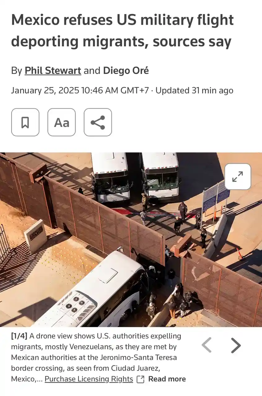An aerial photo shows several white buses parked near a tall border fence, with uniformed personnel and people gathered…