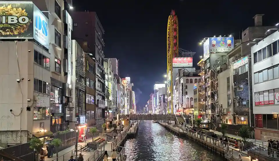 A nighttime photo of a bustling urban canal in Japan, flanked by brightly lit buildings with Japanese signage and a large…