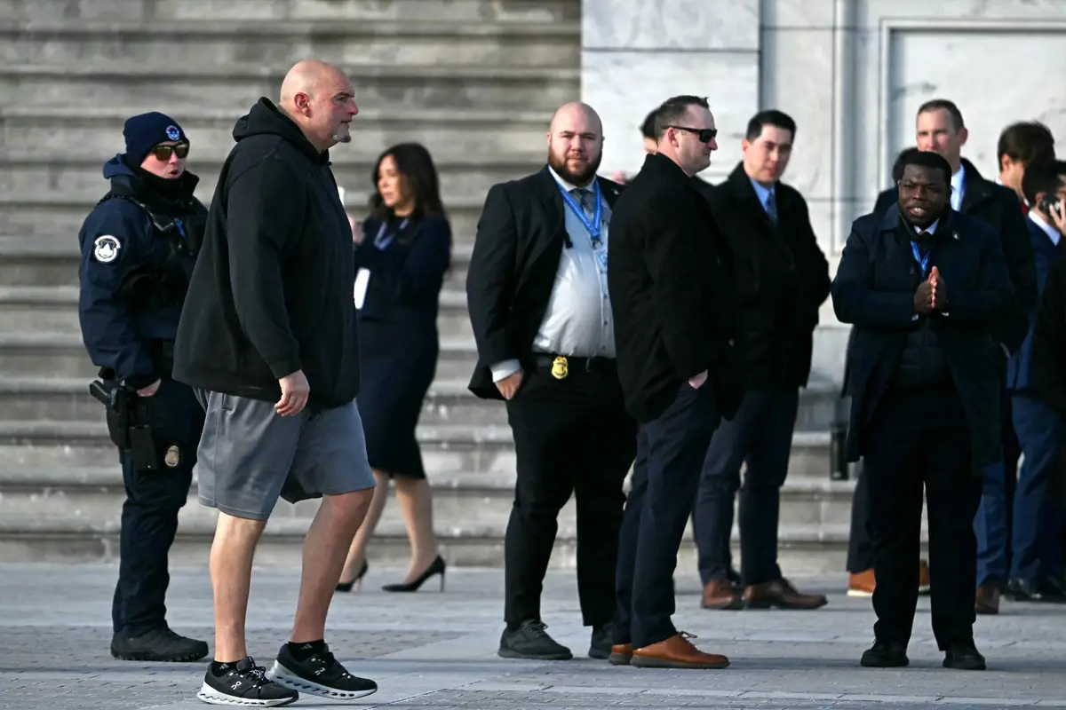 A man in a black hoodie and grey shorts walks past a group of formally dressed people and uniformed security officers…