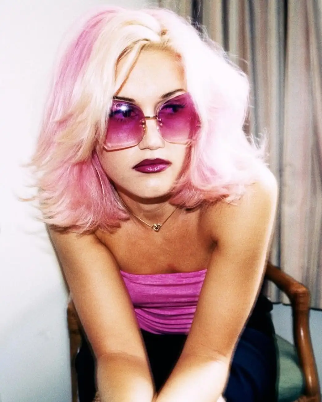 A woman with pink-tipped blonde hair and large pink sunglasses sits in a chair, wearing a pink top and dark lipstick. The…