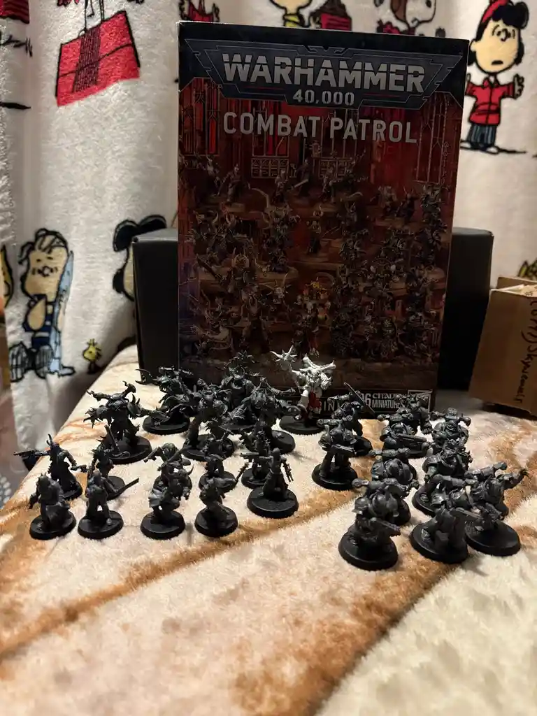 A Warhammer 40,000 'Combat Patrol' box stands behind a large group of unpainted, grey miniature figurines arranged on a…
