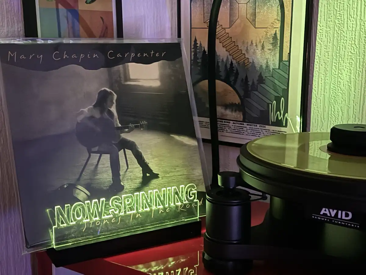 A vinyl record player with a glowing green neon sign that reads 'NOW SPINNING' is positioned next to a Mary Chapin Carpenter…