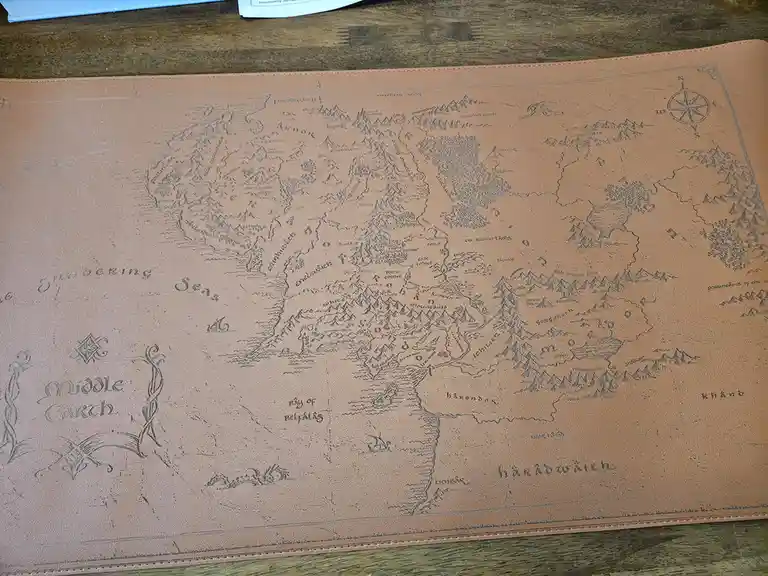 A tan leather desk mat features an embossed map of Middle Earth with detailed fantasy geography, including labeled regions…
