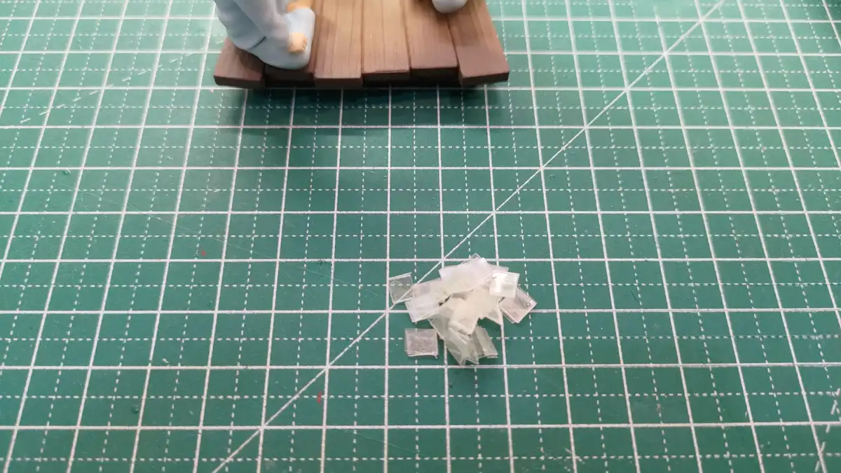 A close-up photo shows a pile of small, shattered glass fragments on a green cutting mat with grid lines. In the upper left…