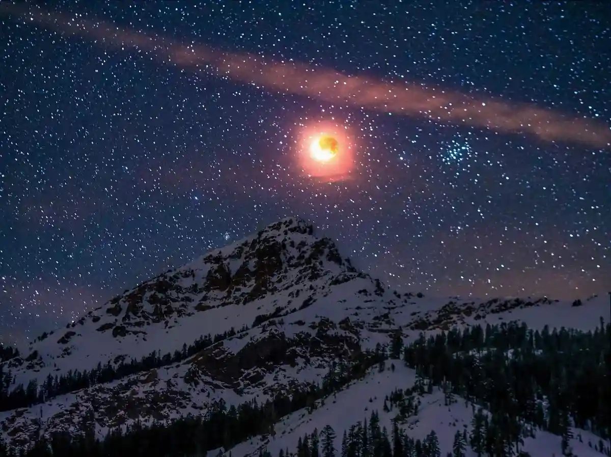 A stunning night sky photo shows a bright, glowing moon partially obscured by a reddish-orange cloud, set against a backdrop…