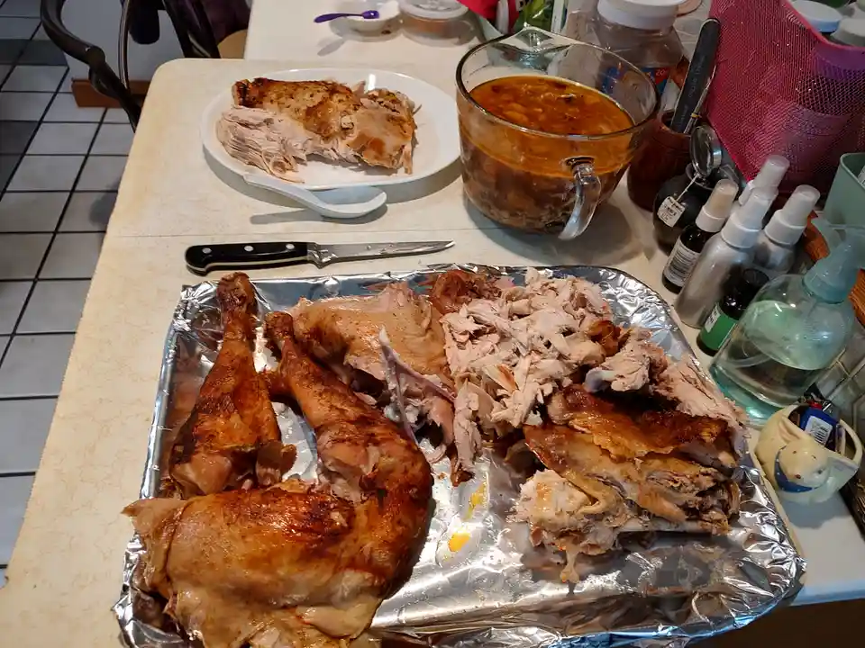 A kitchen counter displays a partially carved roasted turkey on a foil-lined tray, with a plate of shredded meat and a glass…