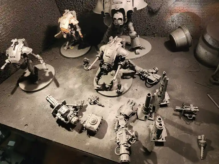 A photo of several large, unpainted sci-fi mech model kits on a textured surface, surrounded by numerous detached weapon…