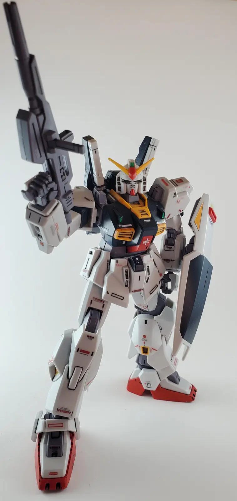 A detailed white, black, and red Gundam model stands in a dynamic pose holding a large rifle and a shield against a plain…