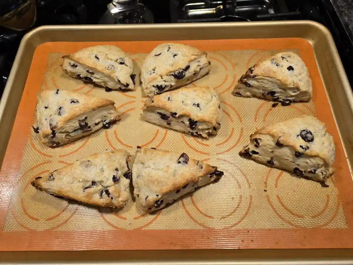 A baking tray holds eight golden-brown triangular scones studded with dark dried fruit, arranged on an orange silicone mat…