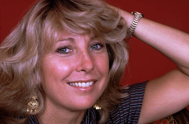 A smiling woman with blonde hair and gold earrings poses against a red background, her hand resting on her head. The…
