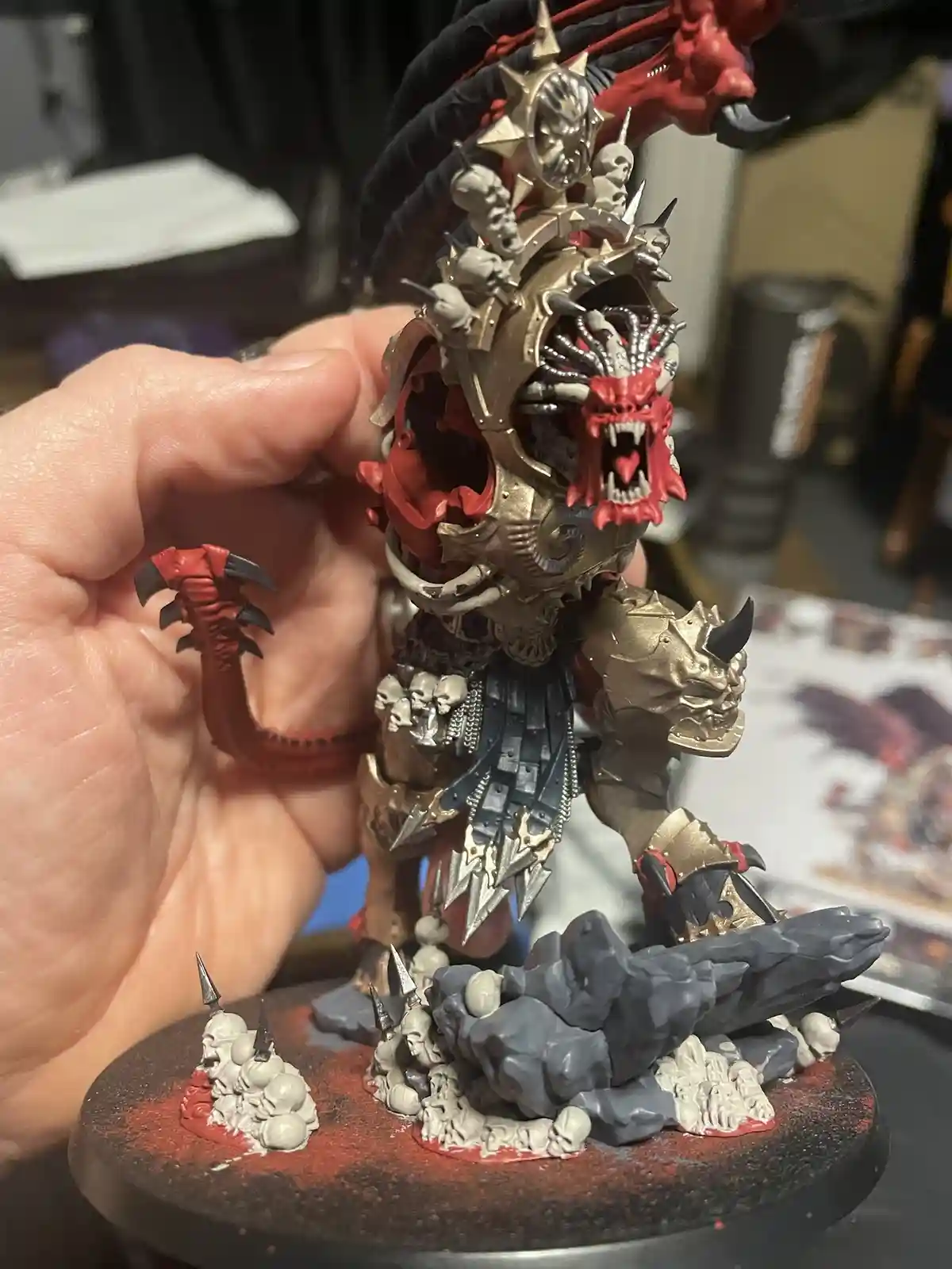 A hand holds a highly detailed, intricately painted miniature of a demonic warlord with red skin, gold armor, skulls, and a…