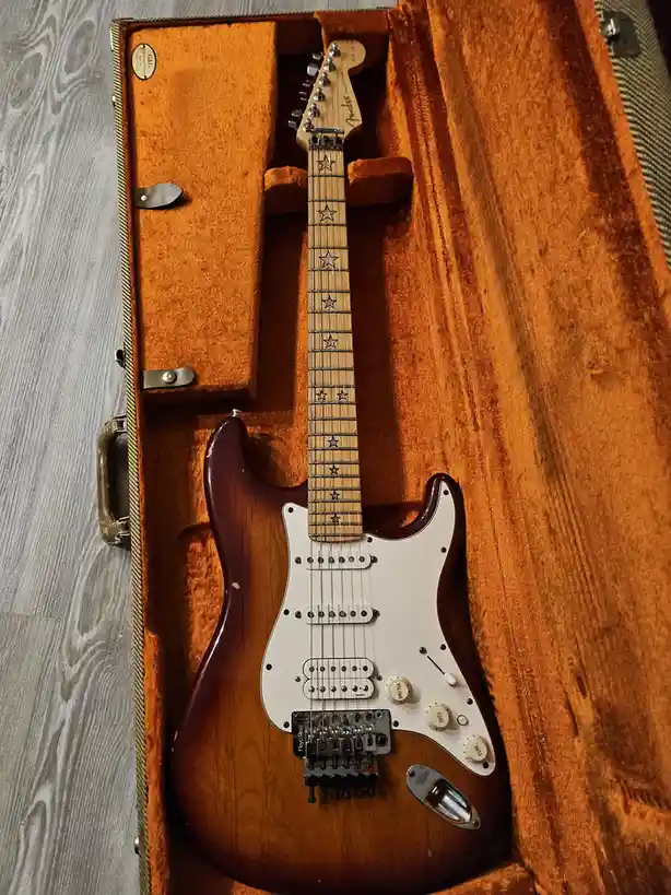 A Fender Stratocaster electric guitar with a sunburst finish and star inlays on the fretboard rests inside its open, orange…