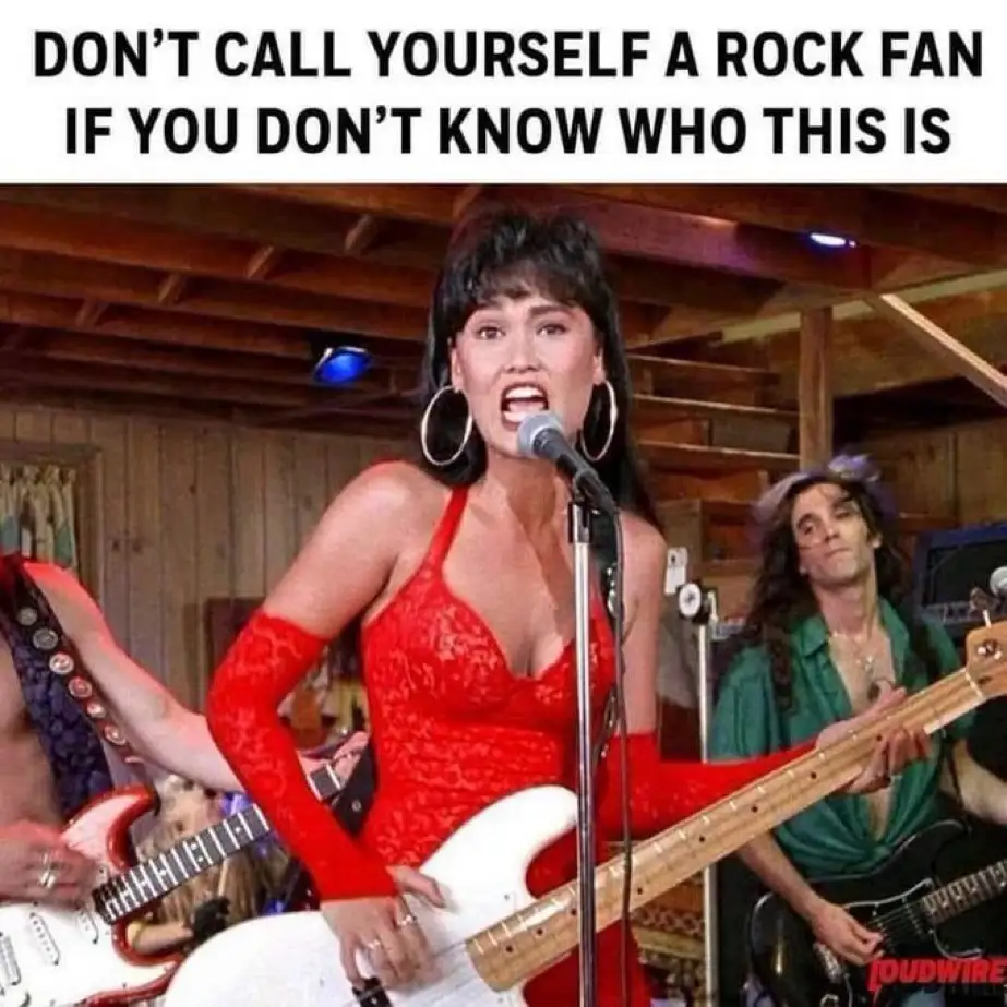 A woman in a red lace outfit and large hoop earrings sings into a microphone while holding a white electric guitar, with two…