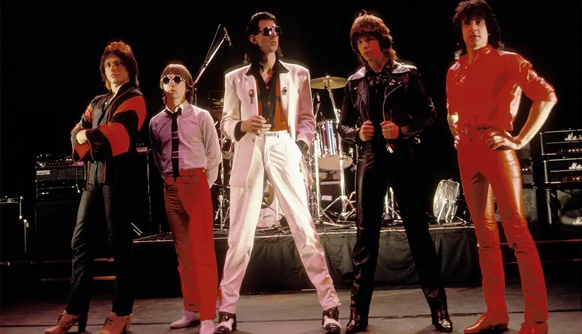 Five men in flamboyant 1970s rock attire stand on a stage with musical equipment behind them. The central figure wears a…