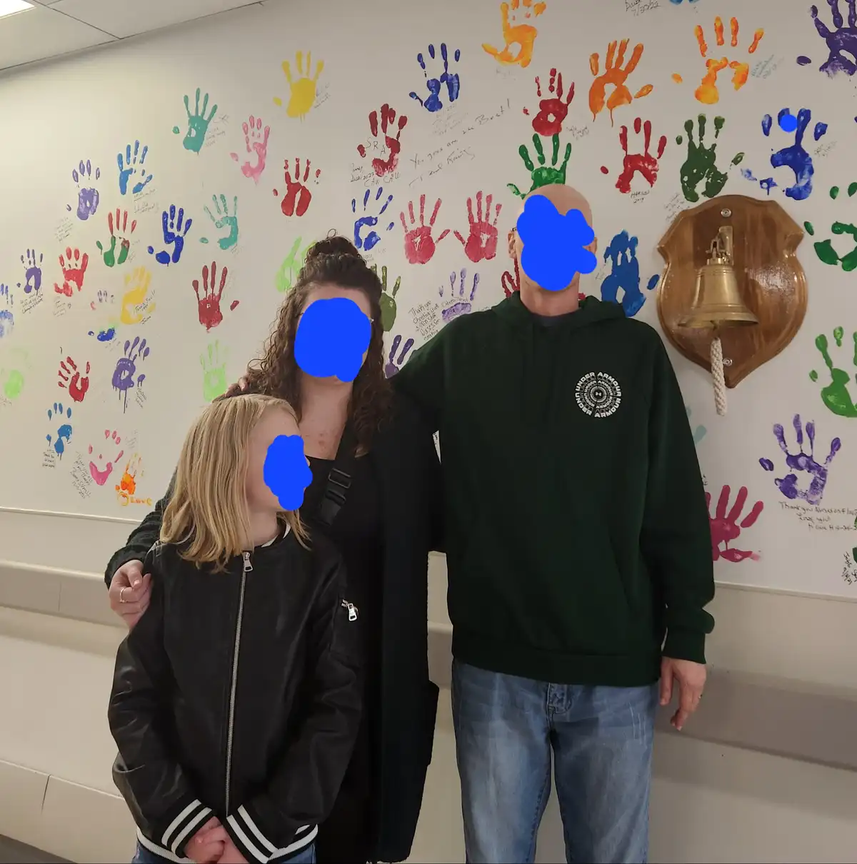 A family of three stands in front of a wall covered in colorful handprints and handwritten messages, likely in a hospital or…