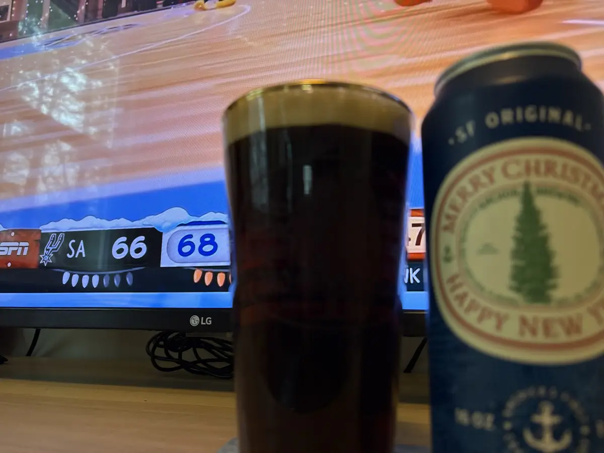 A close-up photo shows a dark beer in a glass and a blue can labeled 'MERRY CHRISTMAS HAPPY NEW YEAR' sitting in front of a…