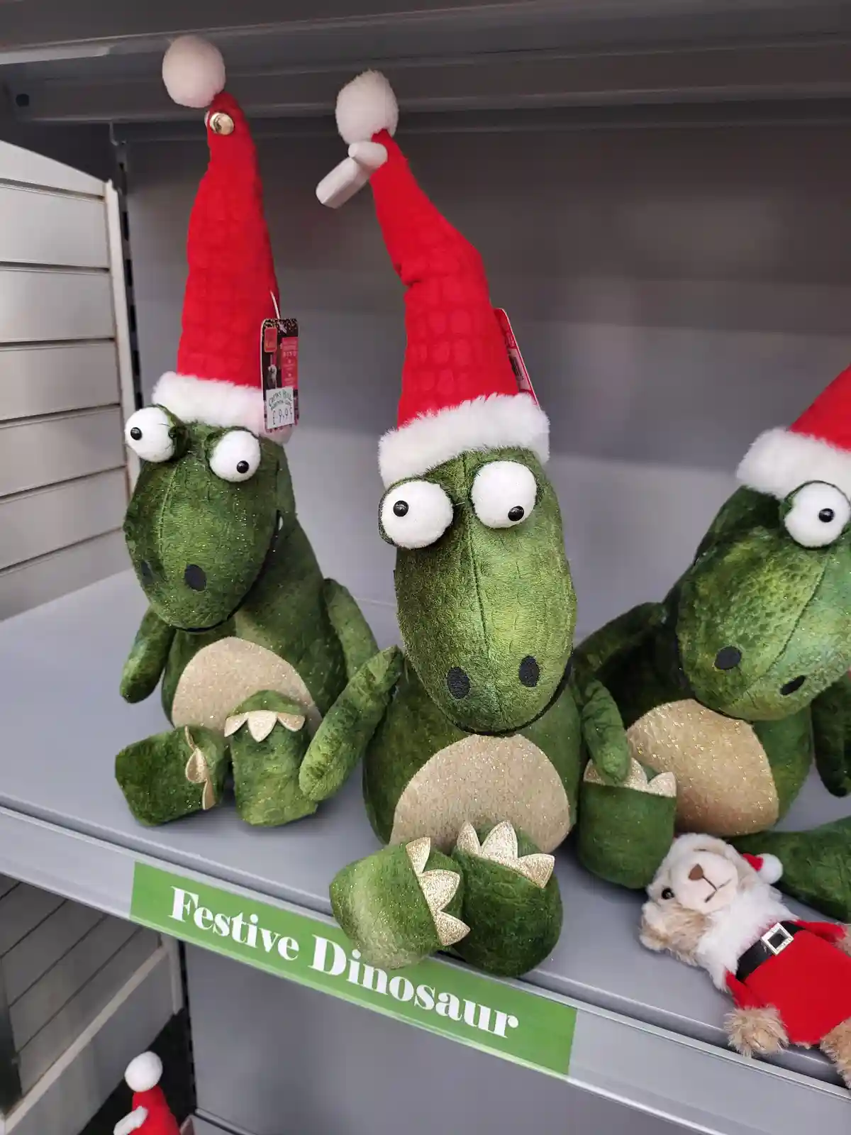 Three green plush dinosaurs wearing red Santa hats sit on a store shelf, with a green sign below them reading 'Festive…