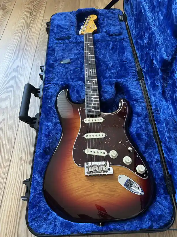 A sunburst-finish electric guitar with a maple neck and rosewood fretboard rests inside an open, black hardshell case lined…
