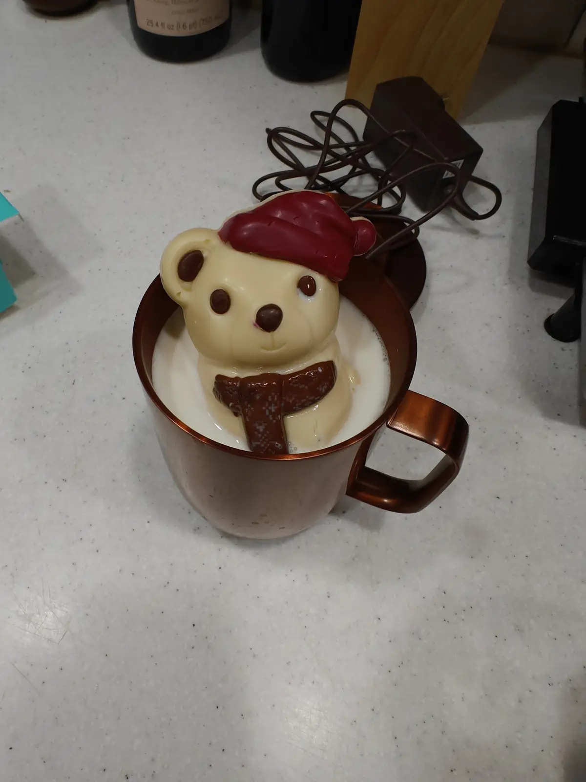 A white chocolate bear figurine wearing a red hat and brown scarf is partially submerged in a copper mug filled with milk…