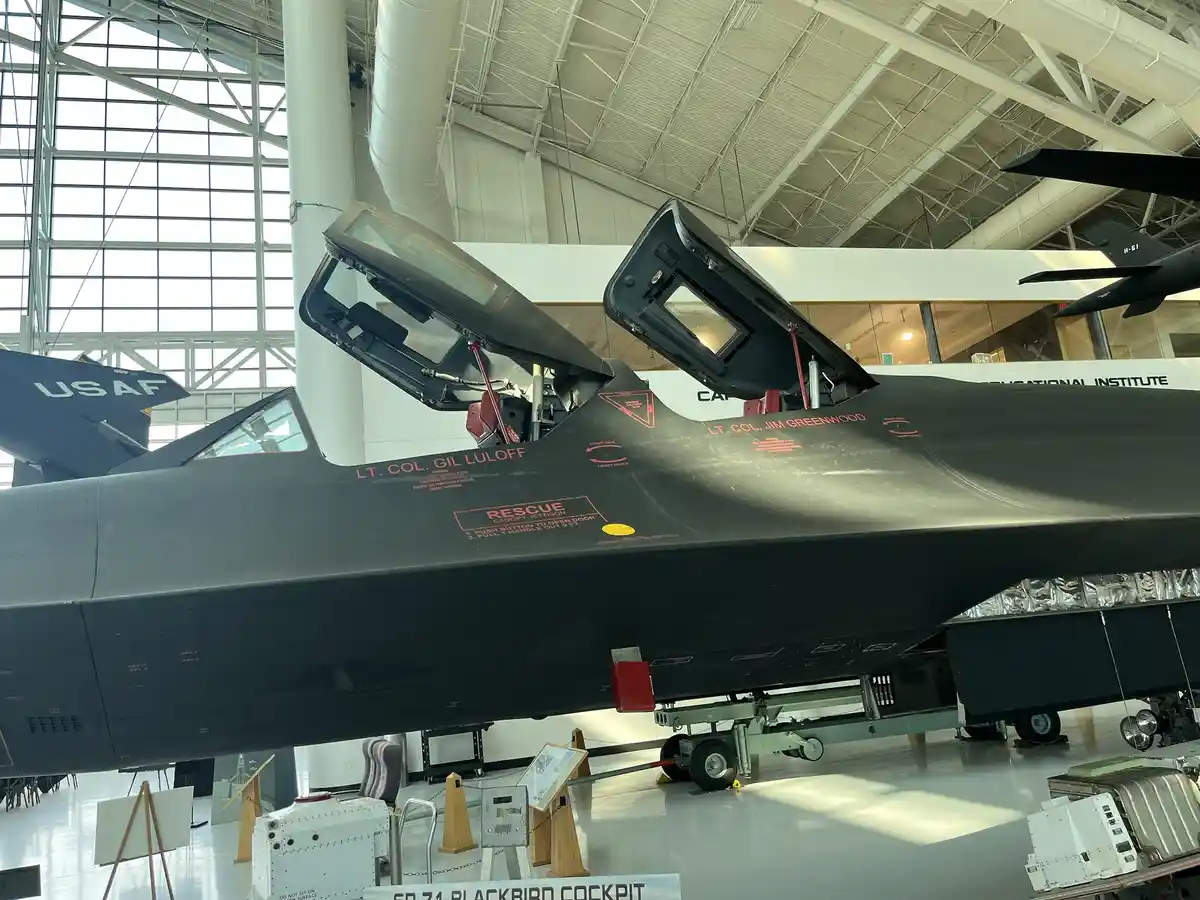 A black SR-71 Blackbird aircraft is displayed indoors with its cockpit canopy open, revealing red text labels including…