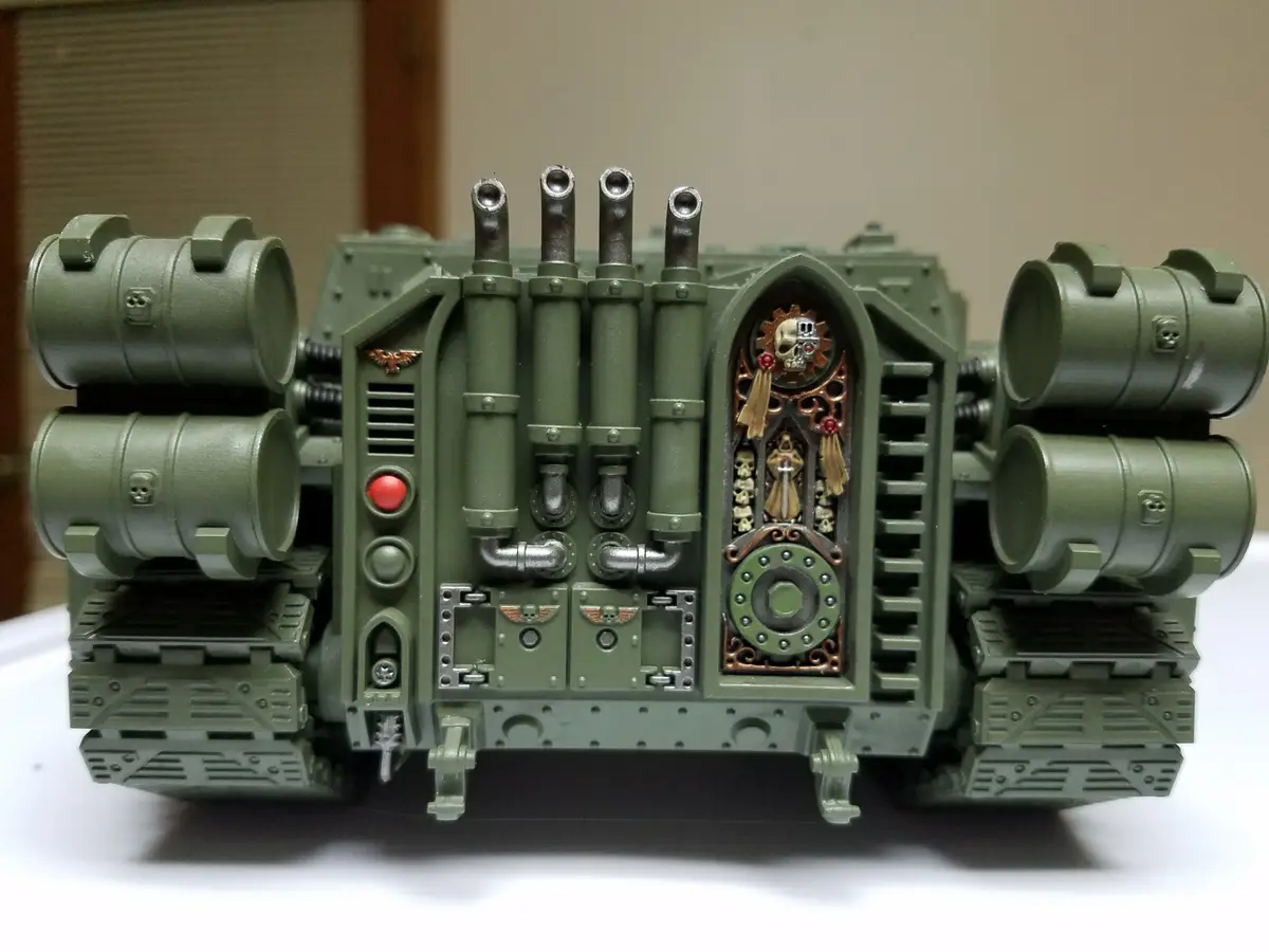 A detailed green Warhammer 40k tank model is shown from the front, featuring four large cylindrical barrels on each side and…