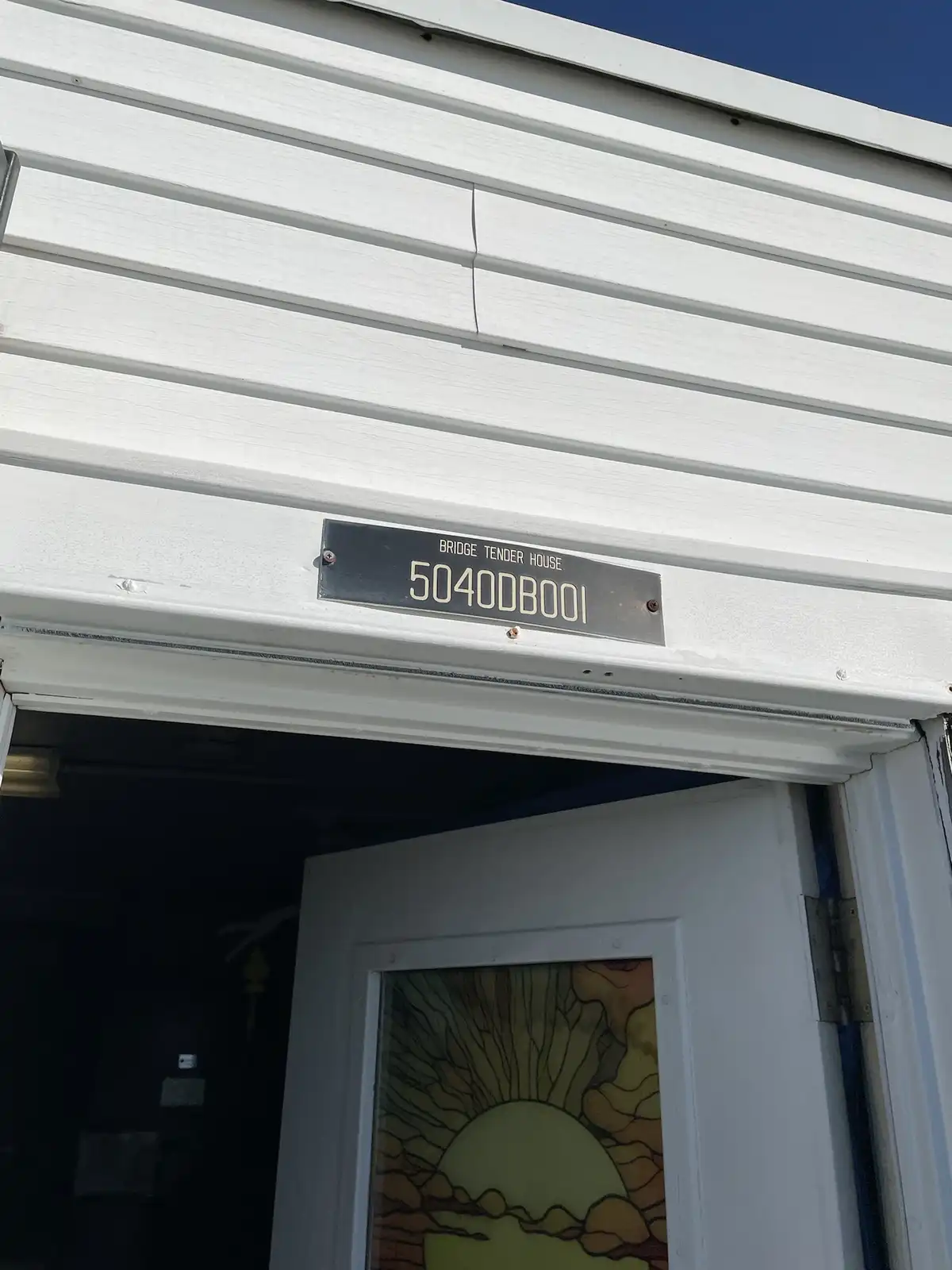 A close-up photo shows a black metal plaque mounted above a white door on a white-sided building. The plaque reads 'BRIDGE…