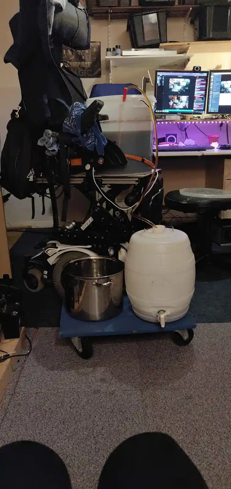 A wheelchair is modified with brewing equipment, including a large white plastic barrel and a metal pot on a blue cart…