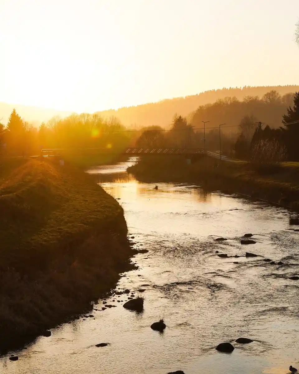 A serene landscape photo of a river at sunrise or sunset, with golden light reflecting on the water, a bridge in the…