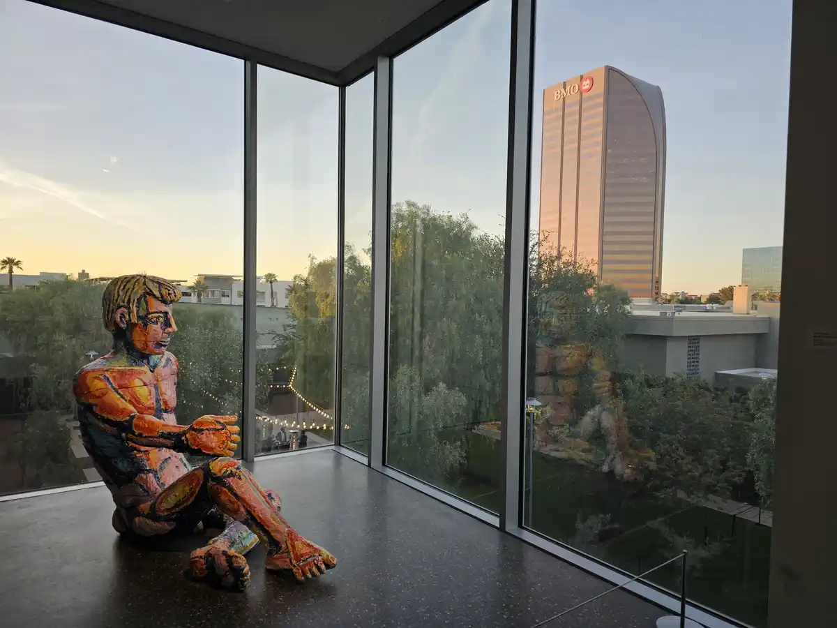 A colorful, stylized sculpture of a seated man is positioned indoors near large windows, looking out at a cityscape that…
