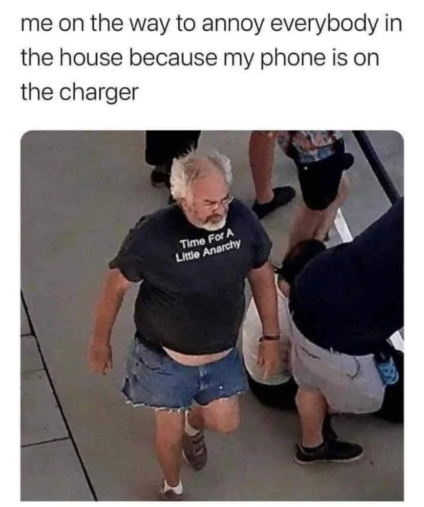 A man with white hair and a beard, wearing a black t-shirt that reads 'Time For A Little Anarchy' and denim shorts, walks…